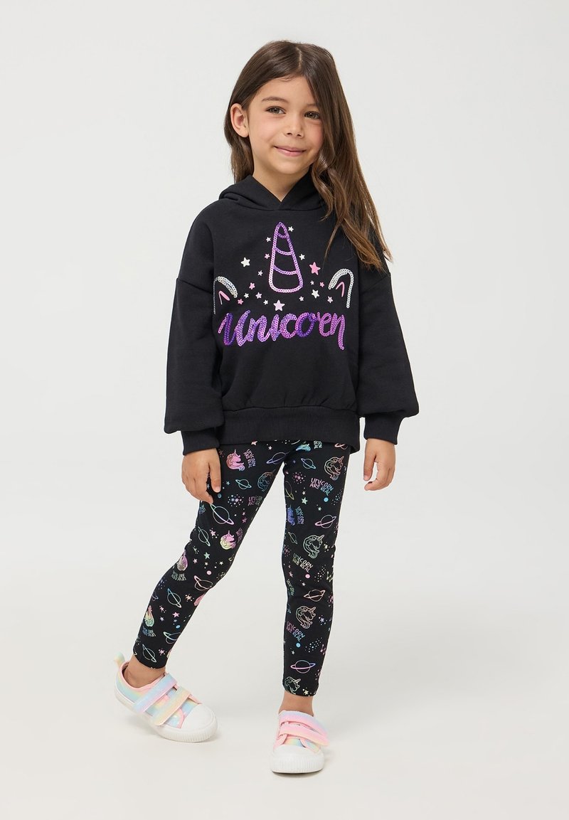 Czarna bluza z kapturem z napisem "Jednorożec" i grafiką, w połączeniu z czarnymi legginsami z kolorowymi wzorami inspirowanymi kosmosem; białe buty z różowymi paskami.