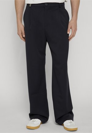 Muž nosi voľné čierne nohavice so stranovými vreckami a biele tenisky s gumovou podrážkou na nevýraznom svetlom pozadí.