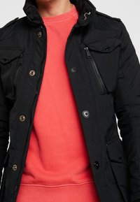 Person som bär en korallfärgad sweatshirt under en ofodrad svart jacka med knappar och dragkedjedetaljer, visad från halsen till midjan.