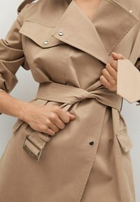 Gros plan sur une personne nouant la ceinture d'un trench-beige avec des boutons en métal et des rabats de poche.