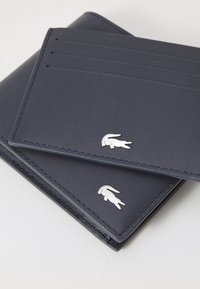 Portofel din piele de culoare bleumarin, cu o textură netedă, dotat cu sloturi pentru carduri și un accent cu logo argintiu pe față. Coaserea pe margini este vizibilă.
