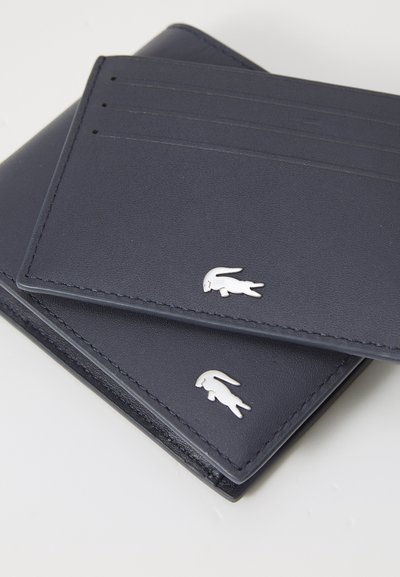 Portefeuille en cuir bleu marine avec une texture lisse, comprenant des emplacements pour cartes et un accent logo en argent sur le devant. Les coutures sur les bords sont visibles.