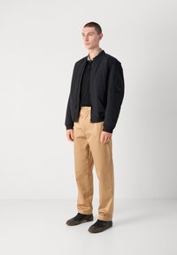 Black lightweight bomber jacket with zip closure, ribbed cuffs, and side pockets, paired with beige trousers and dark shoes.