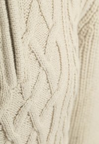 Sweter w kolorze kremowym z warkoczowym splotem, z teksturowanymi wzorami i tkaninowymi detalami oraz ściągaczem przy dekolcie. Miękki, gruby materiał dziany.