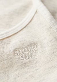 Ljusbeige bomullstyg med en mjuk textur. En vit broderad "Super Dry"-logotyp är synlig på ytan.