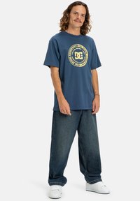 Námořnická modrá bavlněná tričko s kulatým grafickým logem ve žluté barvě, spárované s volnými tmavě modrými džíny a bílými teniskami.