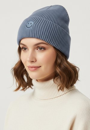 Blue ribbed knit beanie featuring a smiley face patch on the front. The wearer is in a white turtleneck, with soft, light brown hair.