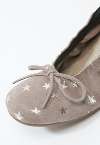 Taupe suède ballerina met een gestrikt strikje en zilveren sterrenpatronen op de teenzone en een lichte elastische afwerking aan de binnenkant van de opening.