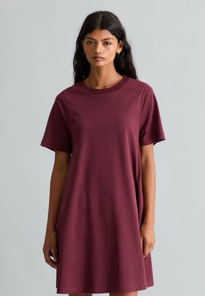 Vrouw met lang donker haar die een loszittende bordeauxrode korte mouw jurk draagt, staand tegen een effen lichtgrijze achtergrond.