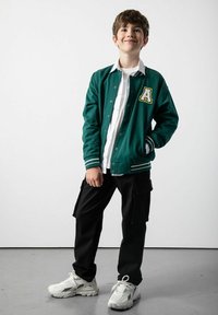 Zielona kurtka akademicka z białymi paskami, wyszywanym literą "A", noszona na białej koszuli; w połączeniu z czarnymi spodniami cargo i białymi sneakersami.