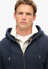 Granatowa bluza z kapturem na zamek błyskawiczny z regulowanym sznurkiem, gładka faktura oraz szary, warstwowy kołnierz z logo. Zamek z przodu z metalowymi akcentami w kolorze srebrnym.