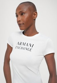 Hvit tettsittende T-skjorte med korte ermer, med svart trykk som sier "ARMANI EXCHANGE" i en fet, stilisert skrift. Glatt bomullstekstur.