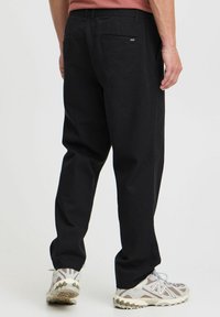 Pantalones negros de algodón con corte recto, con un bolsillo trasero y una pequeña etiqueta de marca. Combinados con zapatillas grises y blancas.