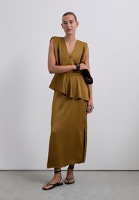 Femme portant une robe sans manches couleur moutarde avec un détail peplum et une fente à l'avant, tenant une pochette noire et portant des sandales à talons noires.