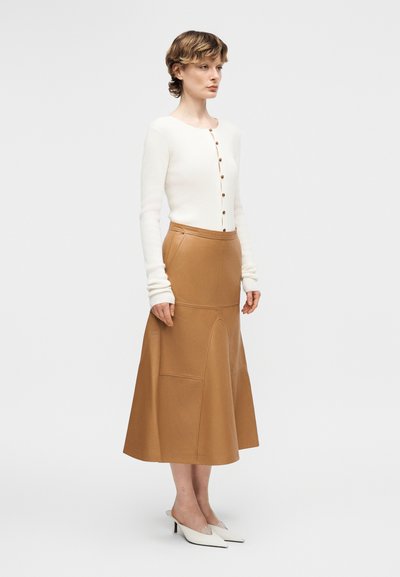 Γυναίκα όρθια, φορώντας λευκό μακρυμάνικο πουκάμισο με κουμπιά, καμηλό midi φούστα σε γραμμή Α με τσέπες και λευκά μυτερά τακούνια σε απλό φόντο.