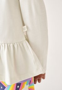 Lichtbeige top met een geplooemde peplumzoom en een klein "Little Bird" label, gecombineerd met levendige, patroonleggings met regenboogkleuren en sterren.