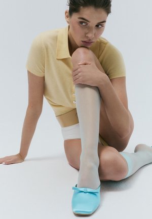 Un haut en tricot jaune clair à manches courtes, des bas légers bleu clair jusqu'aux genoux et des chaussures à enfiler bleues avec un nœud sur un fond clair.