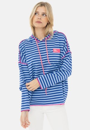 Blonde Frau trägt einen blau-weiß gestreiften Hoodie mit langen Ärmeln, pinken Kordelzügen und Nähten und posiert vor einem einfarbigen Hintergrund.