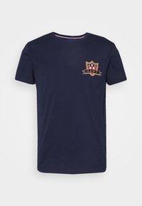 Camiseta de algodón azul marino con cuello redondo, mangas cortas y un logo de escudo bordado con acentos rojos y blancos en el pecho.