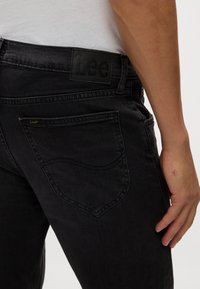 Mustad jeansid, millel on silmatorkav tagatasku ja tekstureeritud vöökoht, millel on tume reljeefne logo. Peen kollane õmblusdetail.