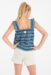 Mėlynai ir baltai dryžuota topas su volanų pečių diržais, su kvadratine nugarėle ir žymės logotipu, derinamas su smėlio spalvos šortais.