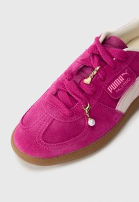 Sneaker Puma Palermo in suede rosa con suola marrone, lacci rosa, charms a forma di cuore dorato e perla, e banda bianca Puma sul lato.