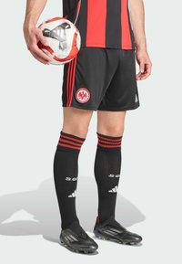 Sort og rød fodboldtrøje med et logo på shortsene, knæ-lange sorte sokker med røde striber og sorte fodboldstøvler, mens han holder en rød og hvid fodbold.