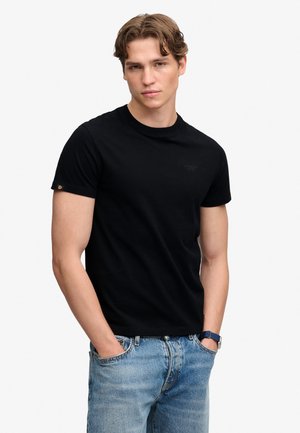 Junger Mann mit kurzen welligen Haaren, trägt ein schwarzes T-Shirt und blaue Jeans, Hände in den Taschen, steht vor einem schlichten weißen Hintergrund.