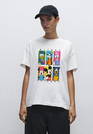 Person iført en hvid T-shirt med farverige Disney-figurer Donald, Goofy, Daisy, Minnie, Mickey og Pluto samt teksten "Mickey og Venner."
