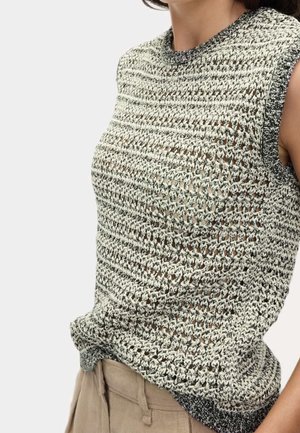 Czarnobiały dziergany top bez rękawów z fakturowanym, ażurowym wzorem, noszony z beżowymi spodniami, ukazujący tors i częściowo szyję.