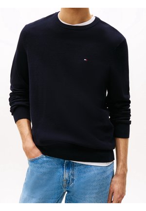 Man draagt een zwart gestructureerd crewneck-trui over een wit overhemd, gecombineerd met lichtblauwe spijkerbroek, hand in de zak, tegen een witte achtergrond.