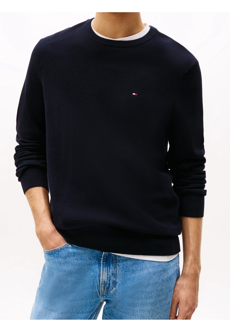 Homme portant un pull ras du cou noir texturé sur une chemise blanche, associé à un jean bleu clair, main dans la poche, sur fond blanc.