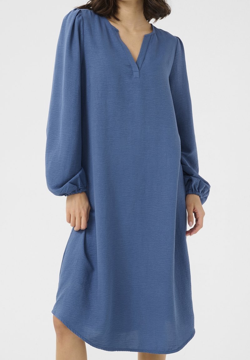 Femme portant une robe bleue ample à manches longues avec des poignets élastiques et un décolleté en V, debout devant un fond uni.
