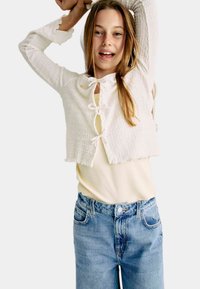 Cárdigan corto blanco con tejido texturizado, cierres frontales anudados y puños con volantes, usado sobre una camiseta amarillo claro y jeans de mezclilla azules.