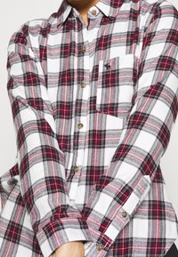 Camicia in flanella a quadri rossi e neri con sfondo bianco, caratterizzata da un design con bottoni, maniche lunghe e una tasca sul petto.