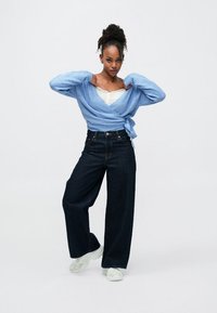 Φόρμα ζακέτας σε ανοιχτό γαλάζιο χρώμα με μακριά μανίκια δεμένη στη μέση, συνδυασμένη με σκουρόχρωμα τζιν φαρδιάς γραμμής και λευκά αθλητικά παπούτσια. Με λεία υφή και άνετη, casual εφαρμογή.