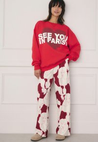 Sweatshirt roșu cu textul „NE VEDEM ÎN PARIS!”, asociat cu pantaloni de pijama cu croială largă, având un model cu pădurica albă și roz.