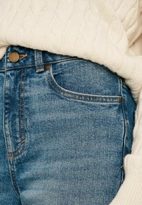 Plave traperice s klasičnim dizajnom s pet džepova, sa prirodnom izblijedjelošću, hardverom u boji bakra i rebrastim krem-svikom iznad.