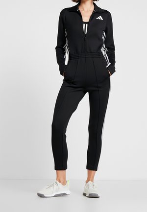 Personne portant une combinaison Adidas zippée noire avec des rayures blanches sur les côtés et des baskets blanches, debout devant un arrière-plan uni.