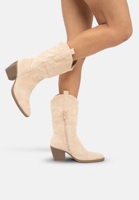Beżowe zamszowe kowbojki z średnią szerokością obcasa, spiczastym noskiem, bocznym zamkiem błyskawicznym i pętelką na górze. Teksturowane wykończenie z subtelnymi detalami przeszyć.