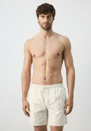 Uomo senza maglietta con capelli corti e ricci che indossa pantaloncini chiari con coulisse, in piedi contro uno sfondo bianco semplice.