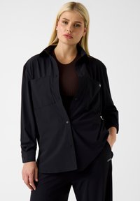 Zwart overhemd met lange mouwen en knoopsluiting, uitgevoerd in een losse pasvorm, met twee borstzakken en een gladde stoffen textuur. Draagt over een donkerribbelige top.