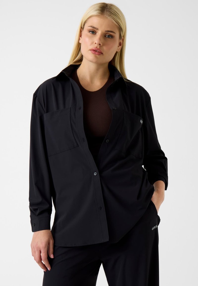 Zwart overhemd met lange mouwen en knoopsluiting, uitgevoerd in een losse pasvorm, met twee borstzakken en een gladde stoffen textuur. Draagt over een donkerribbelige top.