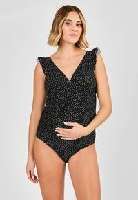 Äitiysuimapuku mustasta kankaasta, jossa on valkoisia pilkkuja, V-pääntie ja röyhelökoristeet olkapäillä. Silkkinen, joustava materiaali.