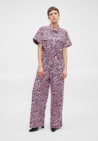 Rózsaszín leopárdmintás overál rövid ujjakkal, széles lábakkal és övvel a deréknál. Fekete gombokkal és fekete kontrasztmintával díszített.