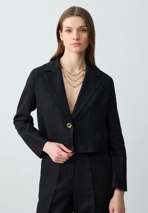 Schwarze, kurze Blazerjacke aus Leinen mit glatter Textur, einem einzigen goldenen Knopf und breiten Revers. Getragen über einem tief ausgeschnittenen Oberteil.