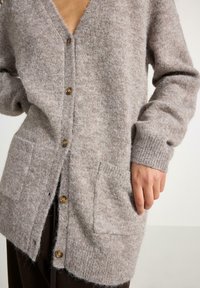 Lysebrun uldcardigan med V-hals, fire gyldne knapper, ribstrikkede manchetter og to forlommer. Blød tekstur og afslappet pasform.