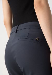 Pantalon bleu marine à texture lisse, avec une poche arrière et un détail en métal doré. Vue rapprochée de la ceinture et du tissu.