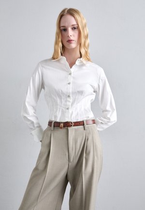 Jeune femme aux cheveux blonds portant une chemise blanche boutonnée rentrée dans un pantalon beige taille haute avec une ceinture marron.