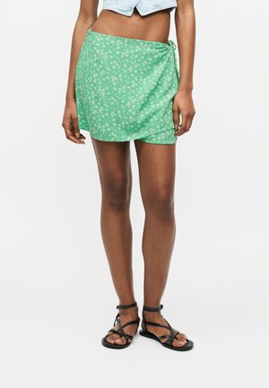 Young person wearing a green floral wrap skirt, cropped light blue denim top, and black strappy flat sandals standing against a plain background.